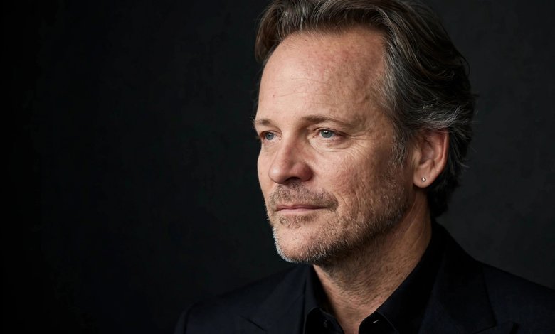Portrait of actor Peter Sarsgaard in a dark suit against a dark studio backdrop.